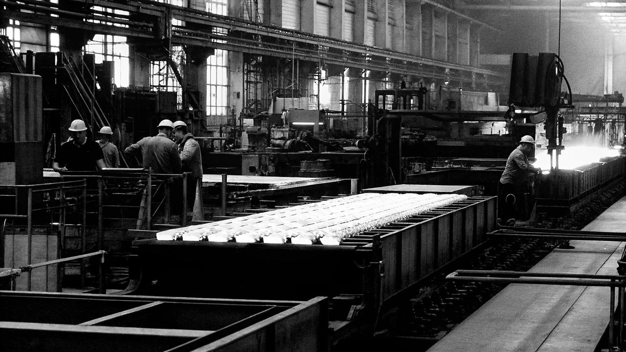 Black-and-white photograph of people in hard hats beside machinery inside a large factory.
