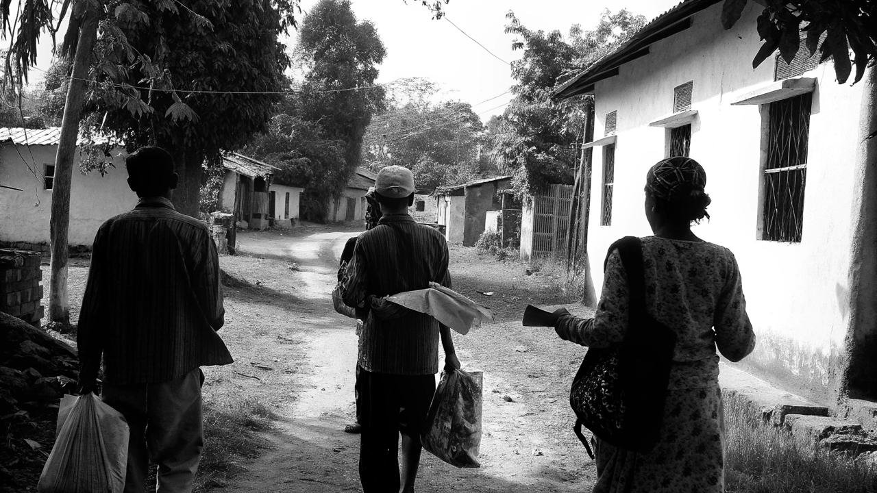 Black-and-white photograph of three people walking along a lane beside buildings and trees.