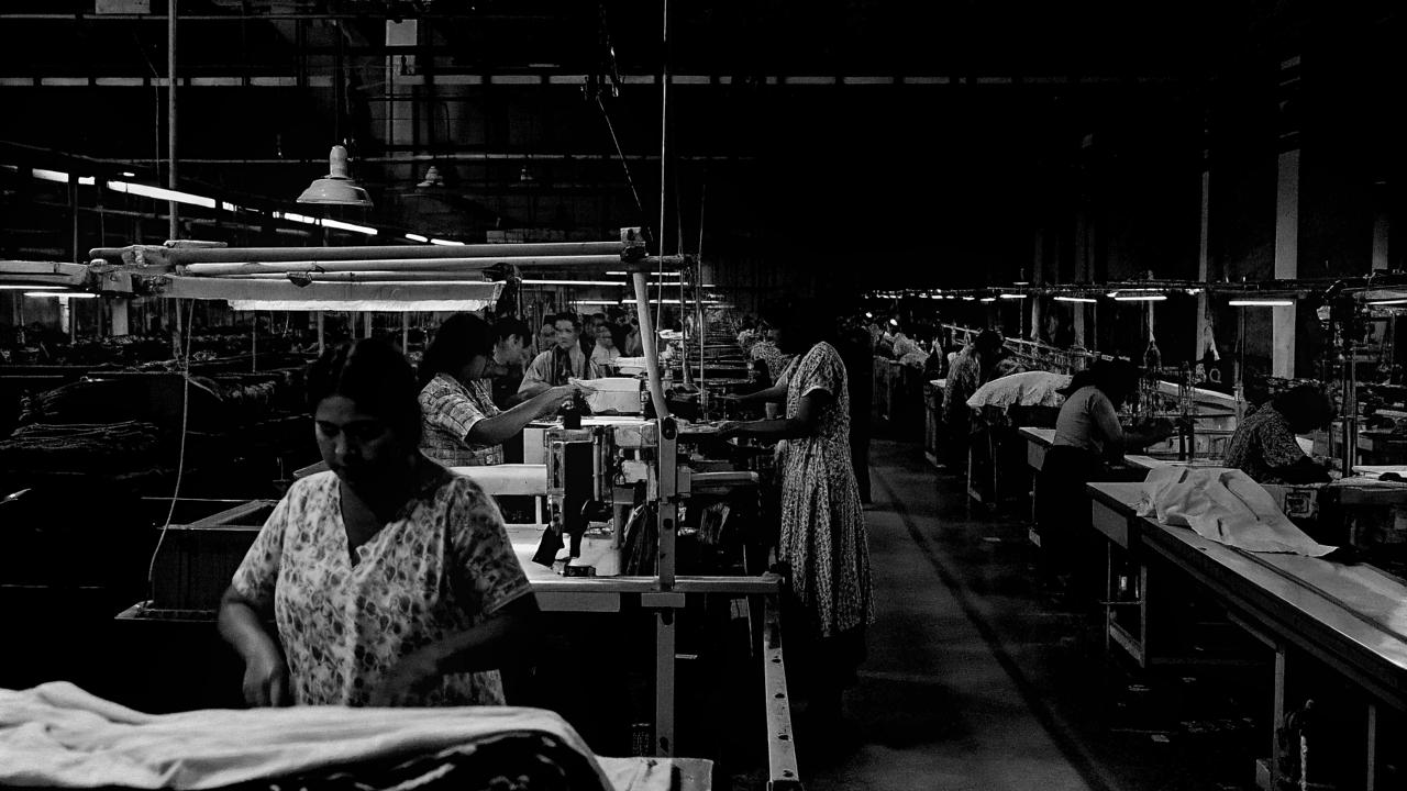 Black-and-white photograph of people working at rows of machines and fabric tables inside a production hall.
