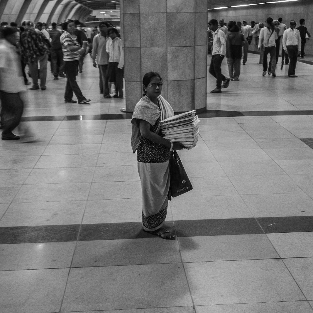 Black-and-white photograph of a woman holding a stack of flat materials in a busy indoor concourse.